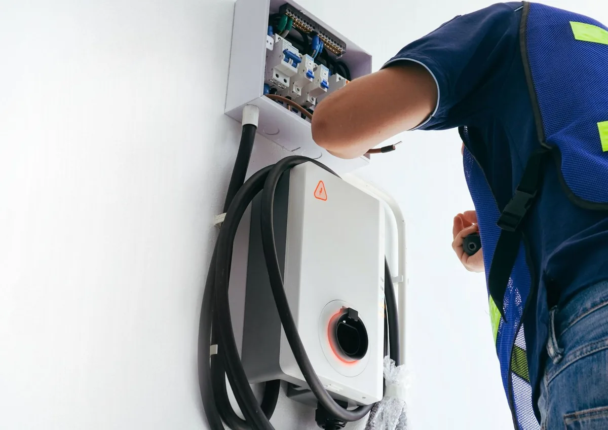 Electrician in a safety vest wiring a small breaker box above a wall-mounted EV charger with its cable coiled